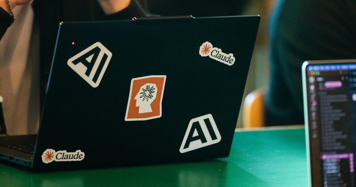 Laptop có sticker Claude và Anthropic trên nắp, đặt trên bàn làm việc lập trình viên với IDE mở trên màn hình