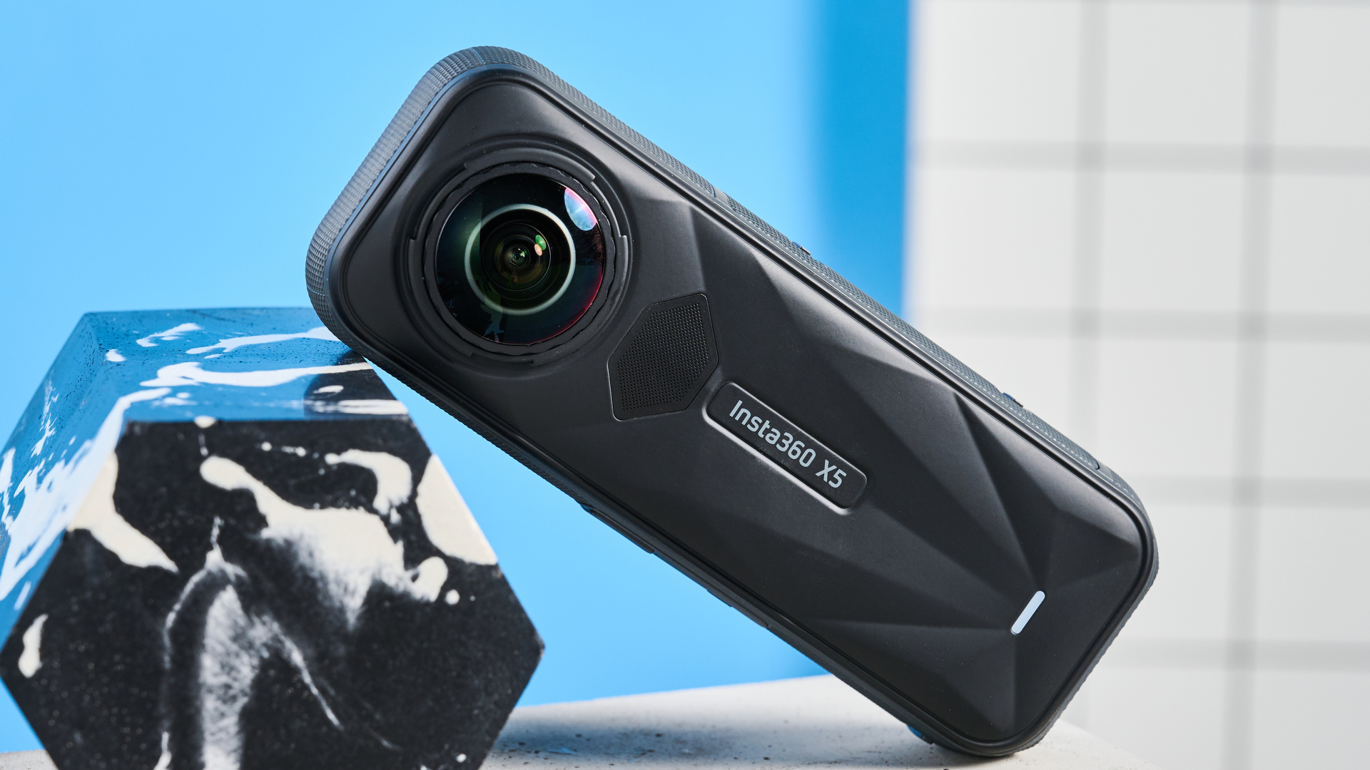 Insta360 X5 camera 360 độ tựa nghiêng cạnh khối đá marble sọc trắng, nền tường xanh